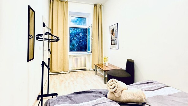 A hotel room with a bed, a chair, a table, a window with curtains, a radiator, and a picture on the wall.