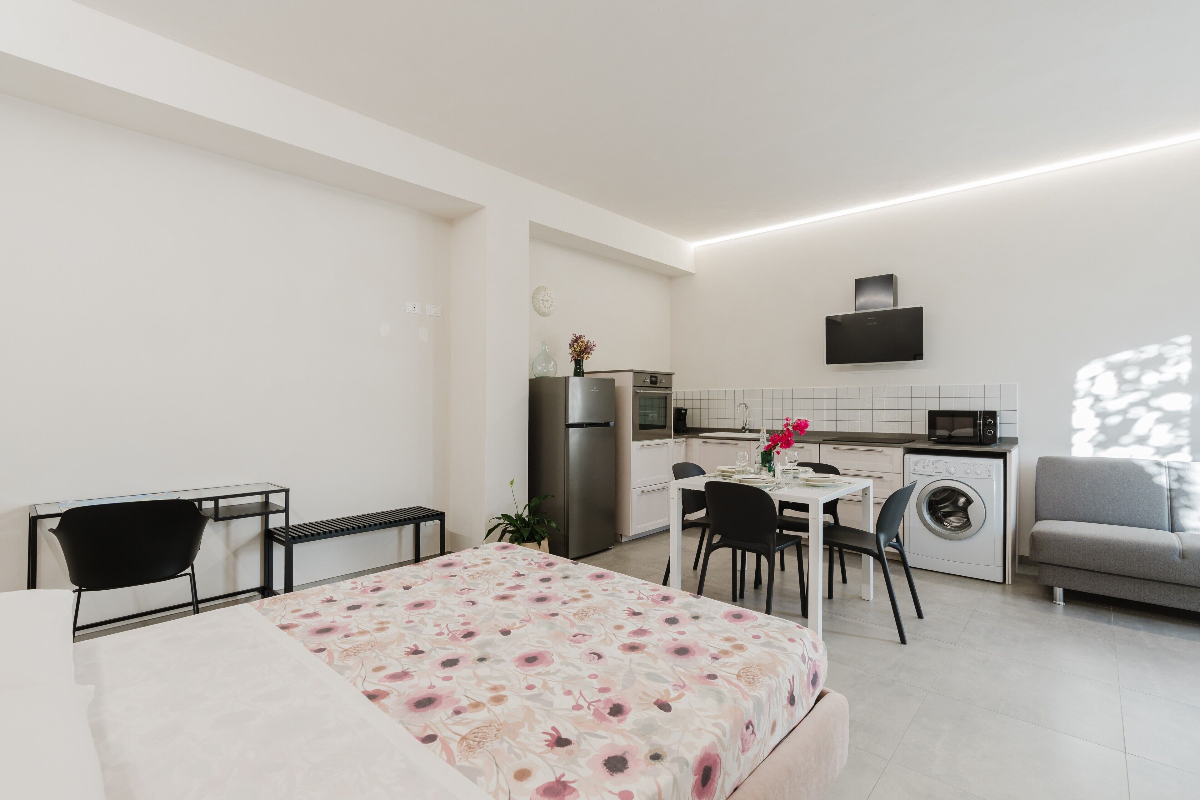 Una habitación moderna y bien iluminada con una cama, una mesa de comedor con sillas, una zona de cocina con microondas, lavadora y un televisor de pantalla plana en la pared.