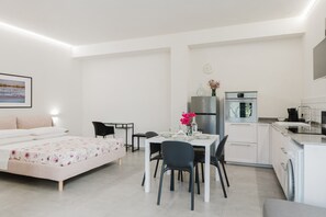 Une pièce moderne et bien éclairée, comprenant un lit, une table à manger avec des chaises, un petit coin cuisine avec un réfrigérateur et un four, et une horloge fixée au mur.