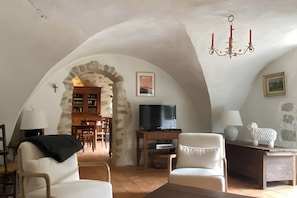 A cozy living room with a TV, white armchairs, a wooden table, and a stone archway leading to a dining area. - House in Provence in a Former Fulling Mill (Entrepierres)