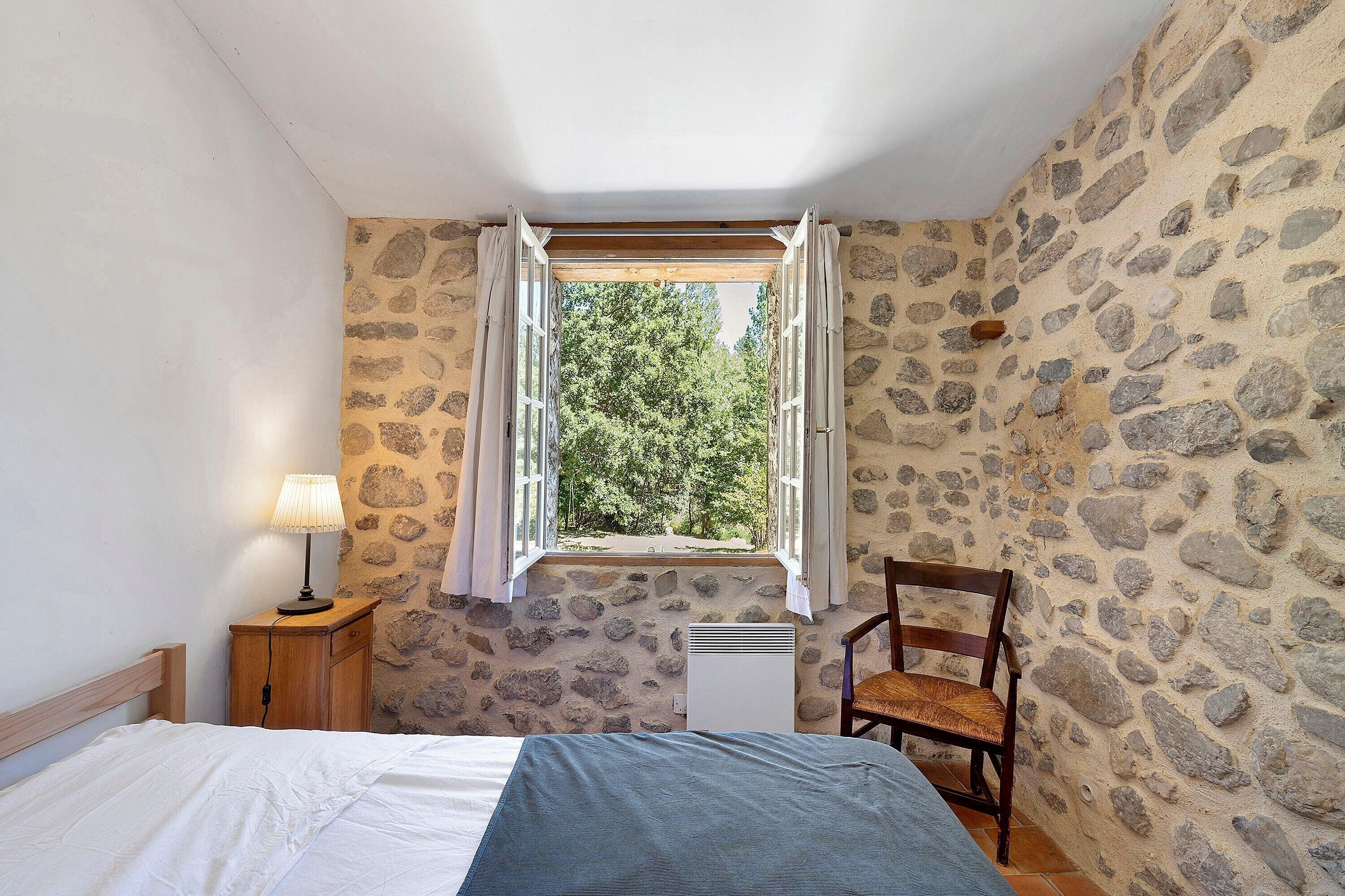 Une chambre avec un mur en pierre, un lit avec une literie blanche et bleue, une table de chevet en bois avec une lampe, une chaise et une fenêtre avec des rideaux.