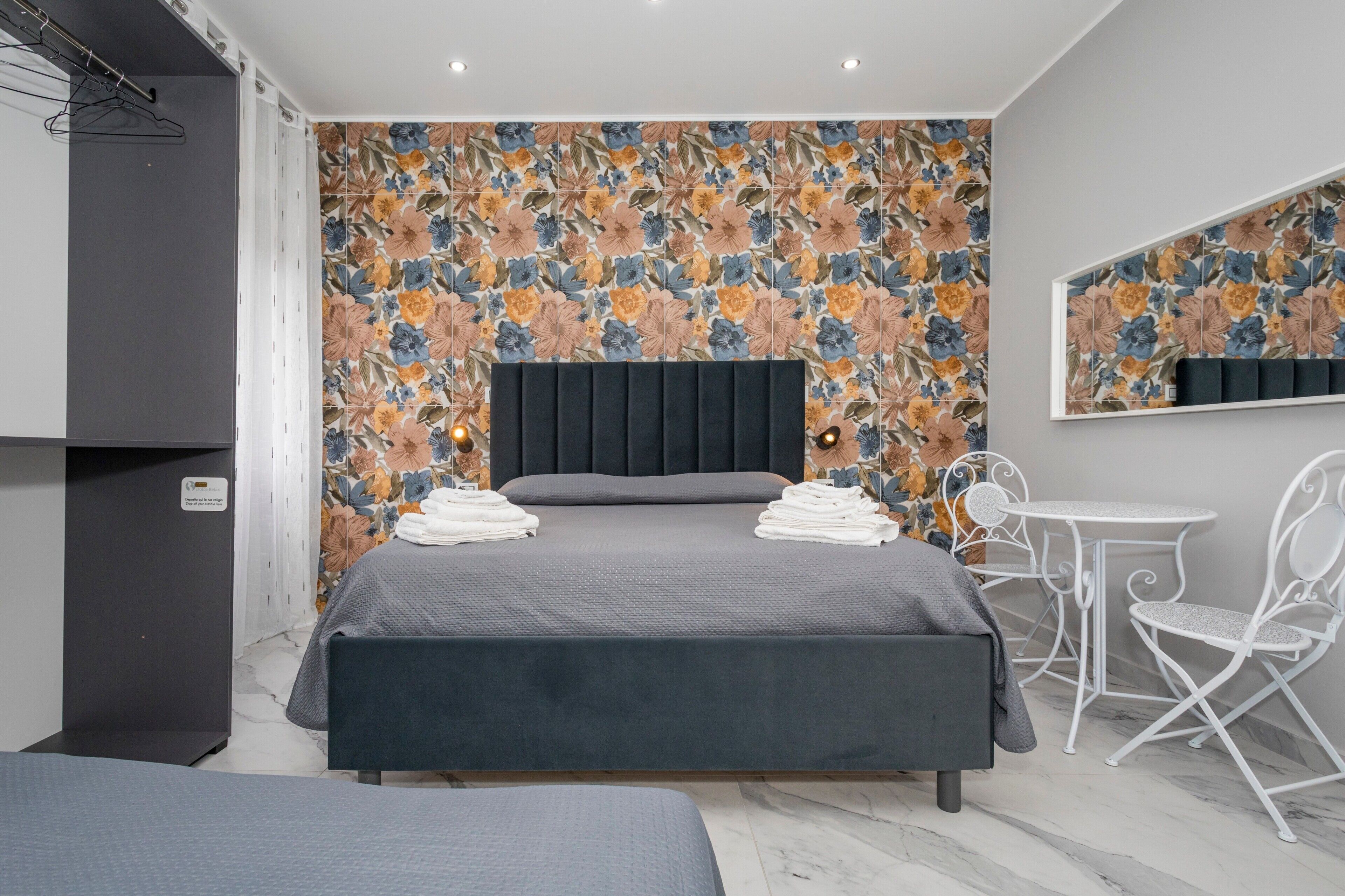 Une chambre d’hôtel moderne avec un grand lit, une petite table et une chaise, une armoire et un mur décoré d’un motif floral.