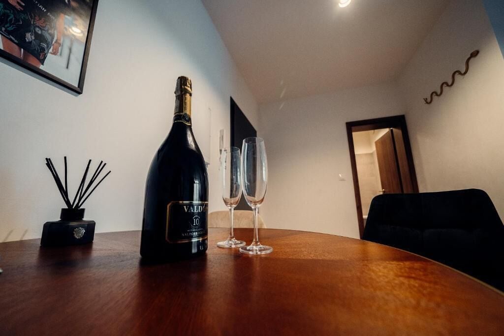 A bottle of Valdeón 10 wine, two wine glasses, and a small black vase with reeds on a wooden table in a room with a dark sofa and a mirror on the wall.