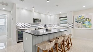 Cocina moderna con armarios blancos, una isla central con taburetes de bar, electrodomésticos de acero inoxidable e iluminación colgante.