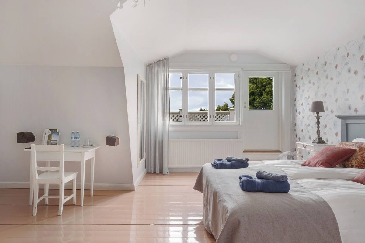 A bedroom with a bed, a desk, and a chair. There is a window with curtains, a radiator, and a lamp on a bedside table.