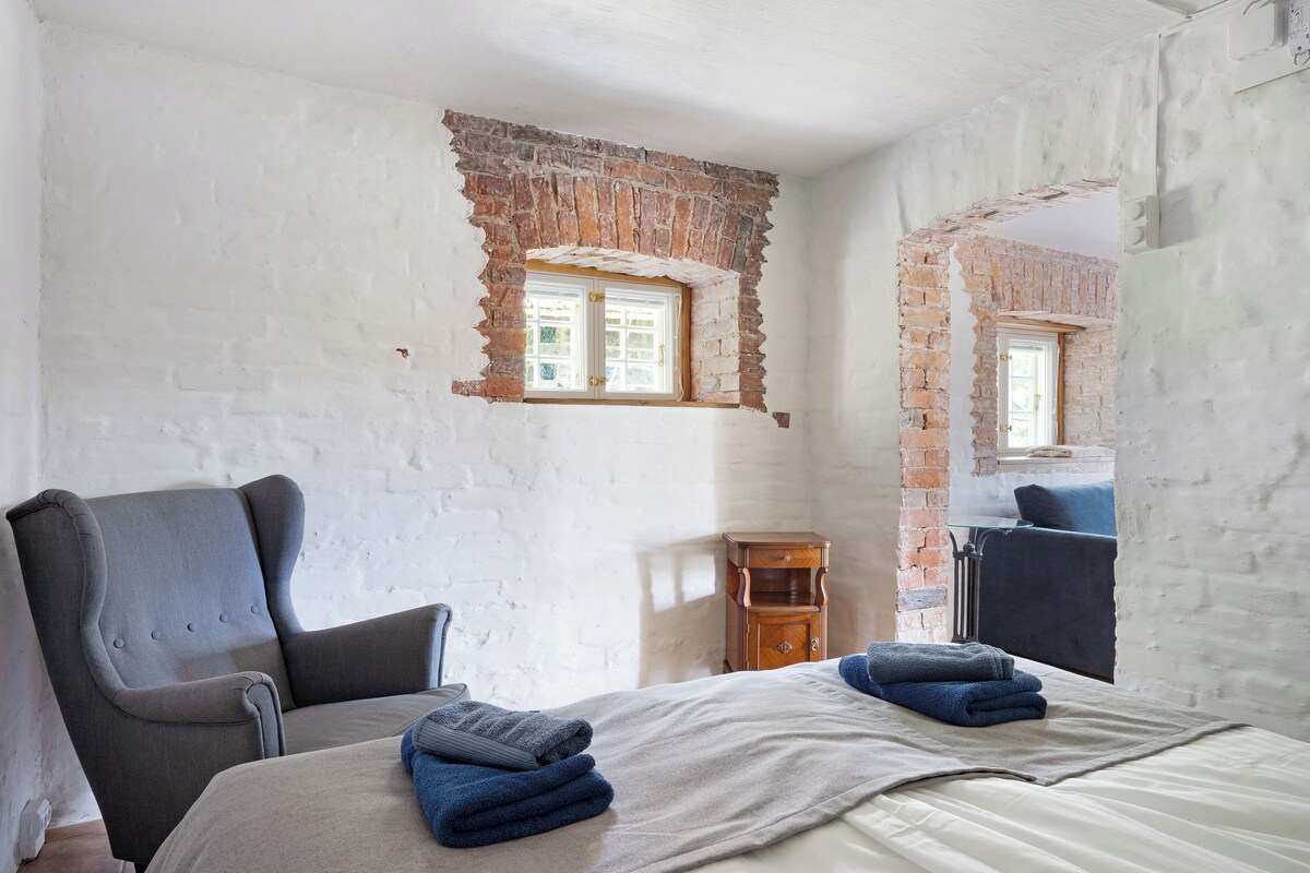 A room with a bed, a chair, a small wooden nightstand, and a window with a brick wall.