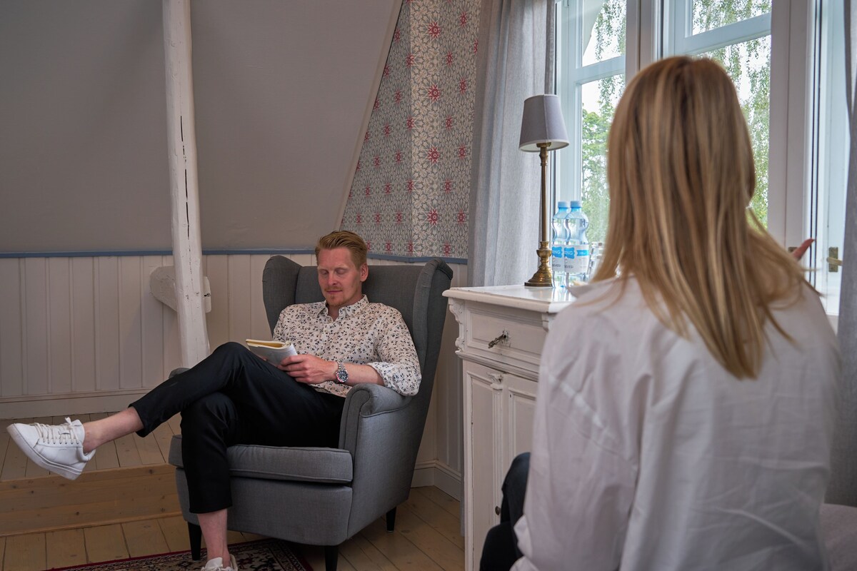 A man sitting in a grey armchair in a room with a patterned wall and a window.