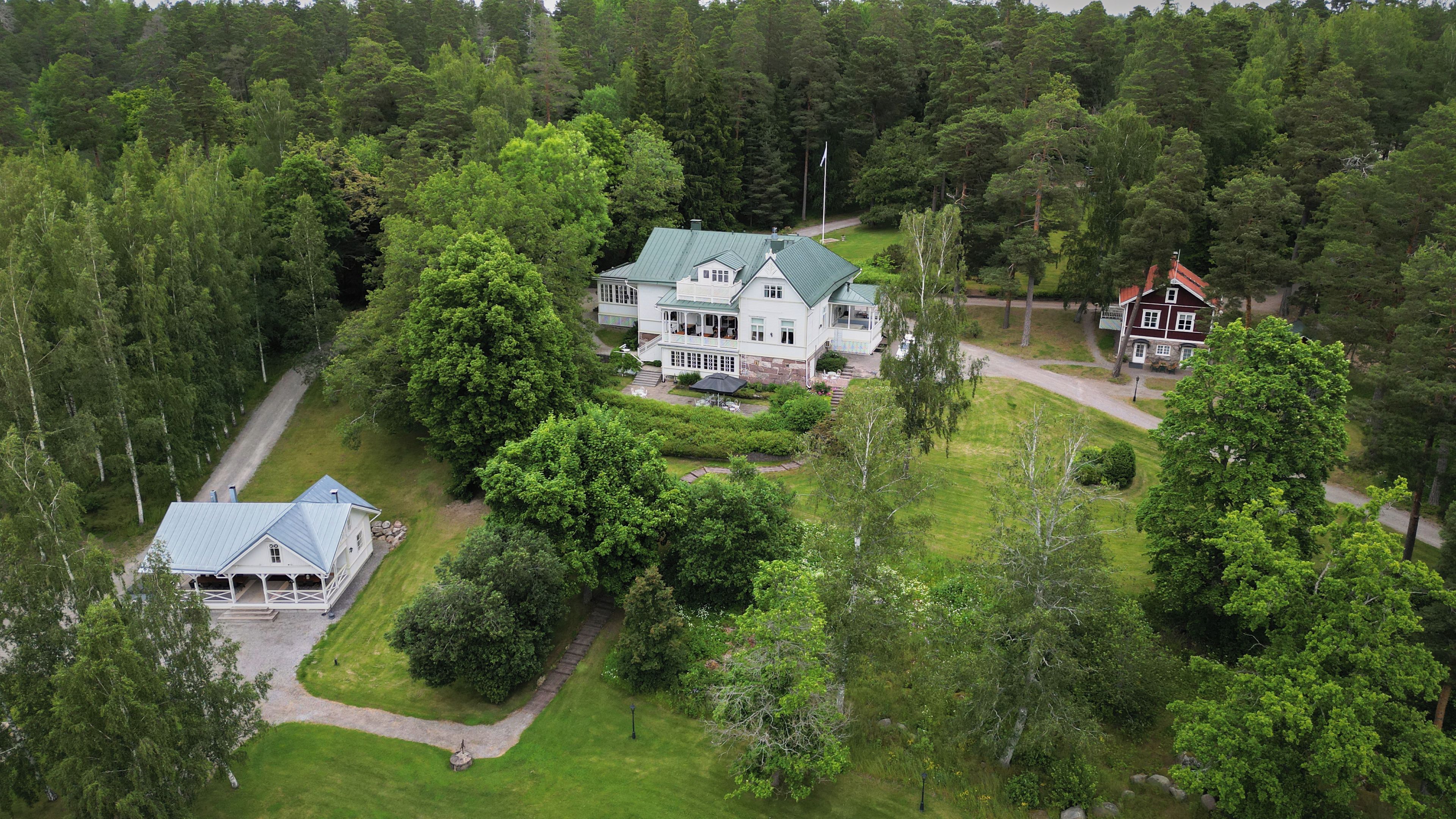 A large, two-story house with a green roof, surrounded by dense forest.