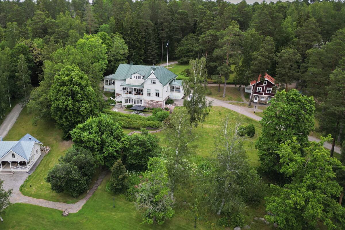 A large, two-story house with a green roof, surrounded by dense forest.