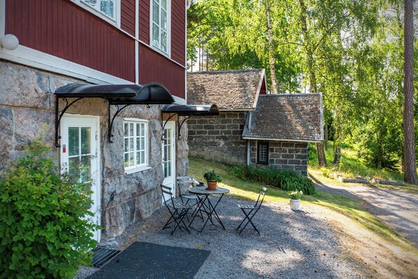 A stone building with a red exterior, a small stone structure, and a gravel patio with a table and chairs. - Villa Wolax (Kaarina)