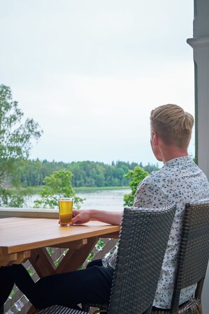 Una persona sentada a una mesa de madera con un vaso de jugo de naranja, con vista a un lago y árboles al fondo.