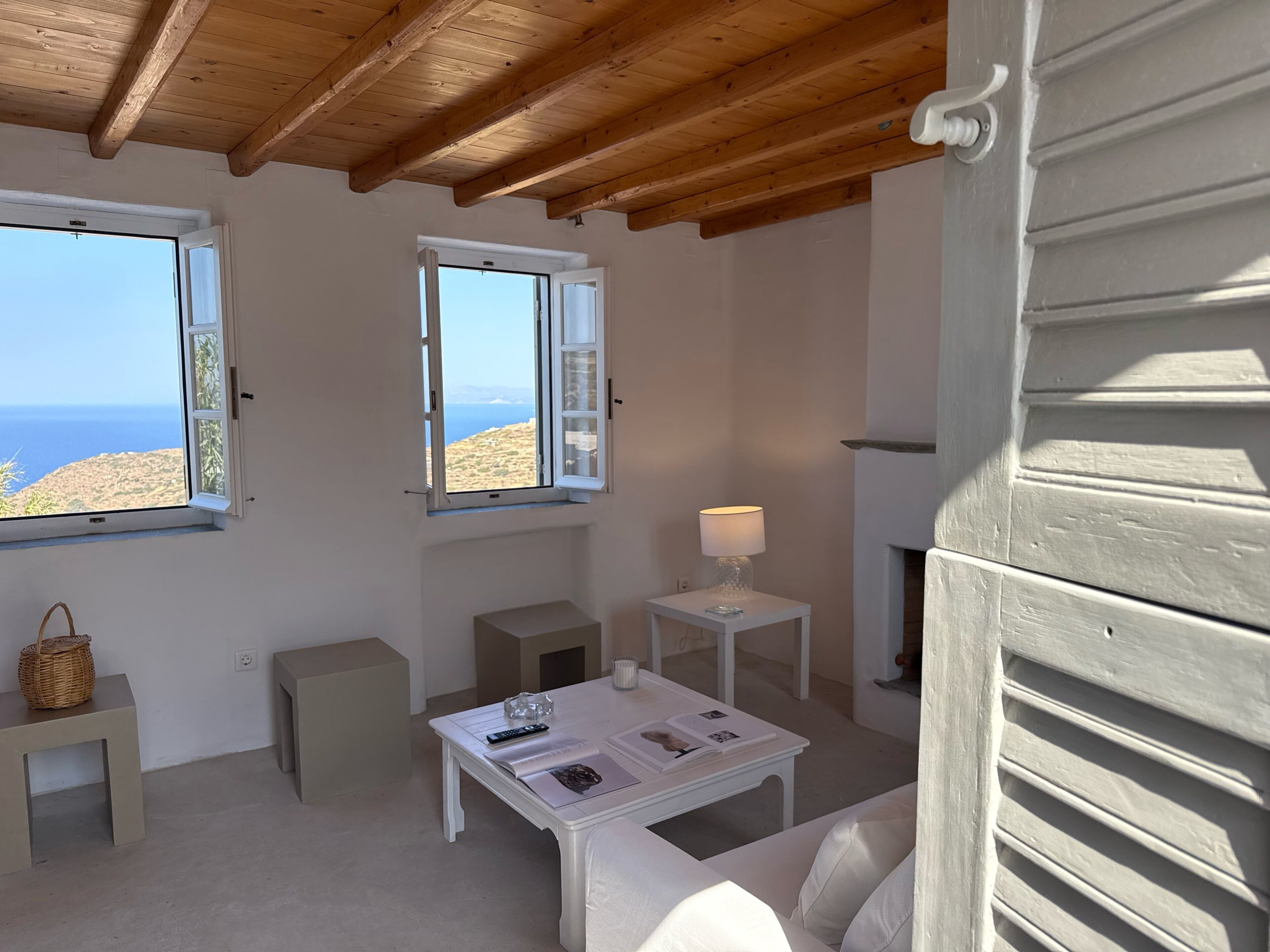 Ein Zimmer mit Holzbalkendecke, zwei Fenstern mit Meerblick, einem weißen Tisch mit Lampe und einem weißen Sofa.