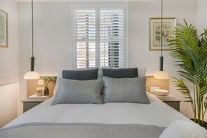 A bedroom with a bed, two bedside tables, a window with blinds, a hanging light fixture, and a potted plant.