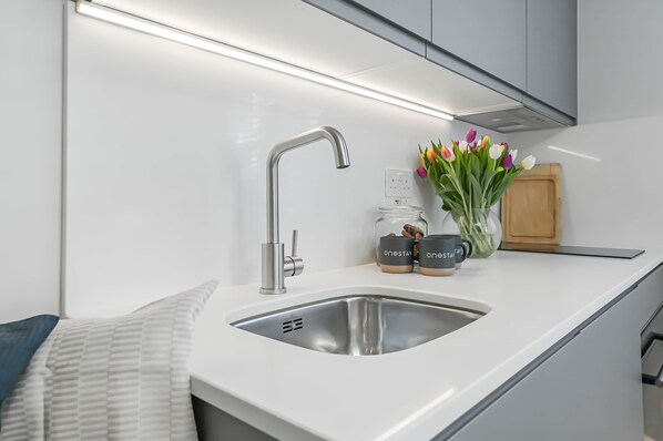 A modern kitchen with a stainless steel sink, a faucet, and a countertop. There are two jars with the text 'ONESTAY' on them, a vase with tulips, and a wooden cutting board.