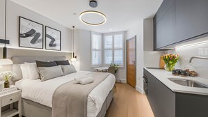 Ein modernes Hotelzimmer mit Bett, Nachttisch und einer kleinen Schreibtischzeile. Ein Fenster mit Jalousien, eine Küchenzeile mit Spüle und Schränken sowie eine Deckenleuchte sind vorhanden.