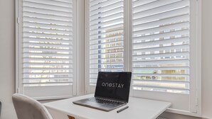 A laptop on a white table with a chair, near a window with blinds.