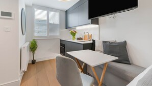A modern kitchenette with a built-in oven, a small dining table, and a grey sofa. - Studio 12 @ OneStay Maida Vale (London)