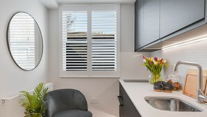 A modern kitchen with a built-in sink, a window with blinds, a mirror, and a potted plant. - Studio 12 @ OneStay Maida Vale (London)