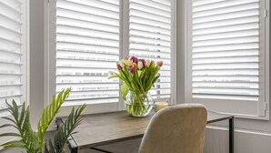 A room with a desk, a chair, a vase with flowers, and a window with blinds.