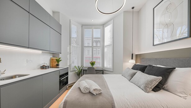 A modern hotel room with a bed, bedside table, and a view of the city through large windows.