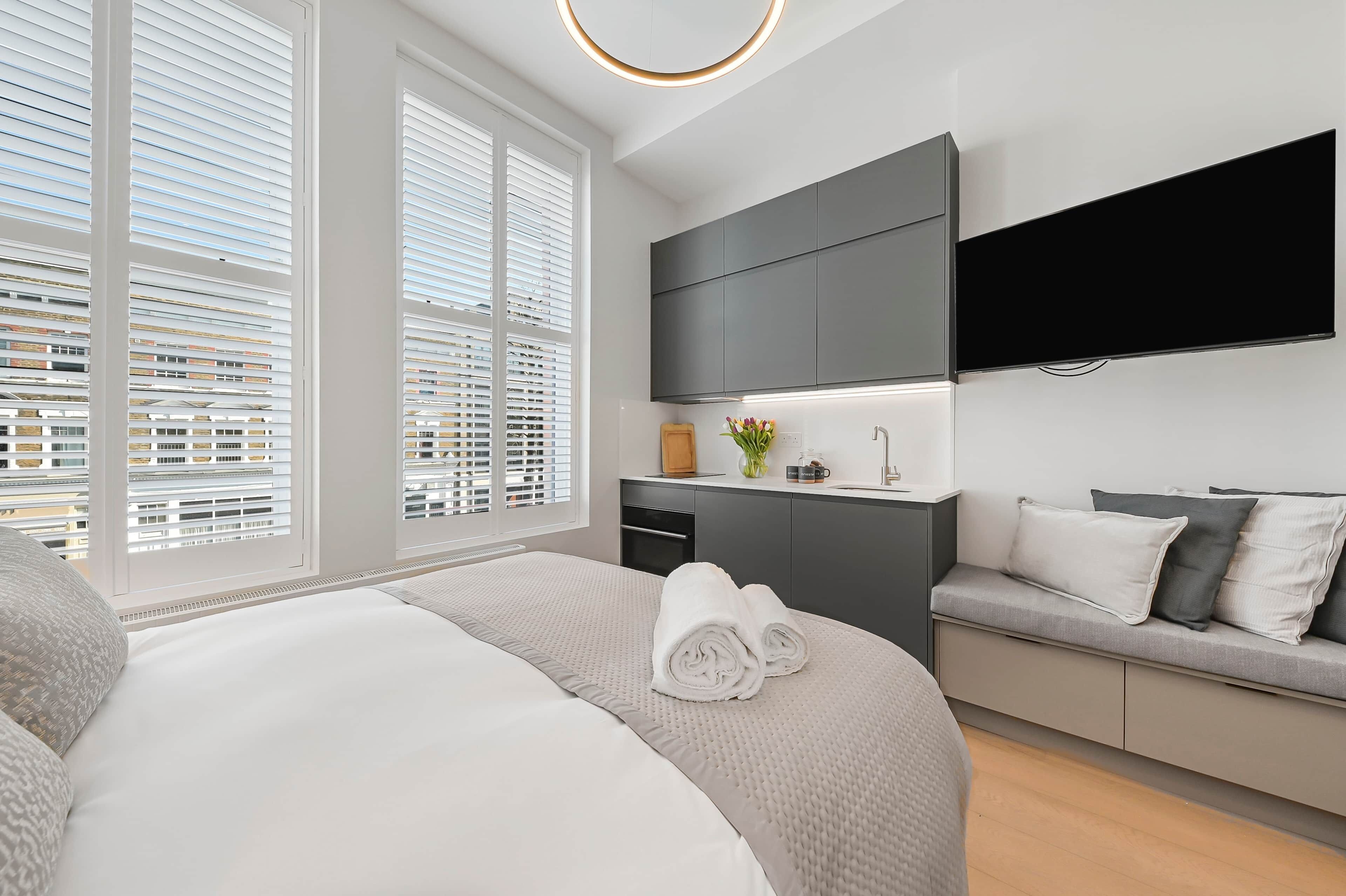 A modern hotel room with a large bed, a sofa, a flat-screen TV, and a view of the city through large windows.
