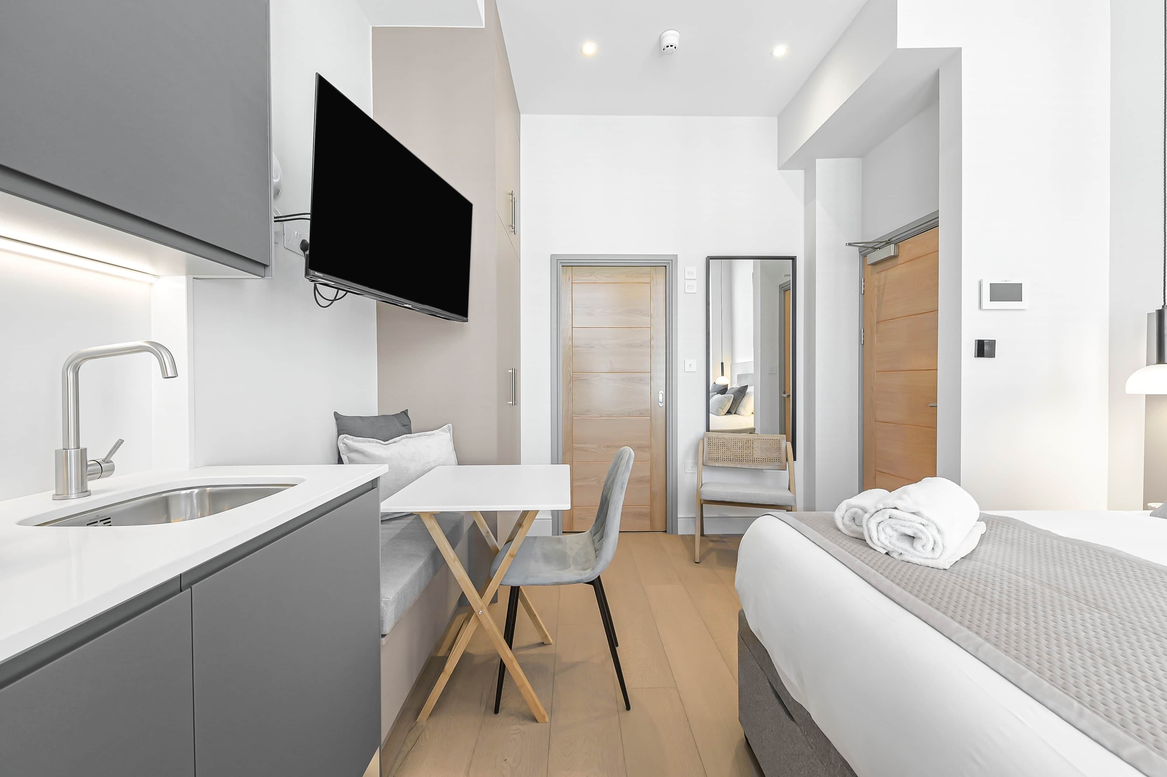 A modern hotel room with a kitchenette, a flat-screen TV, a bed with a rolled towel, and a mirror.
