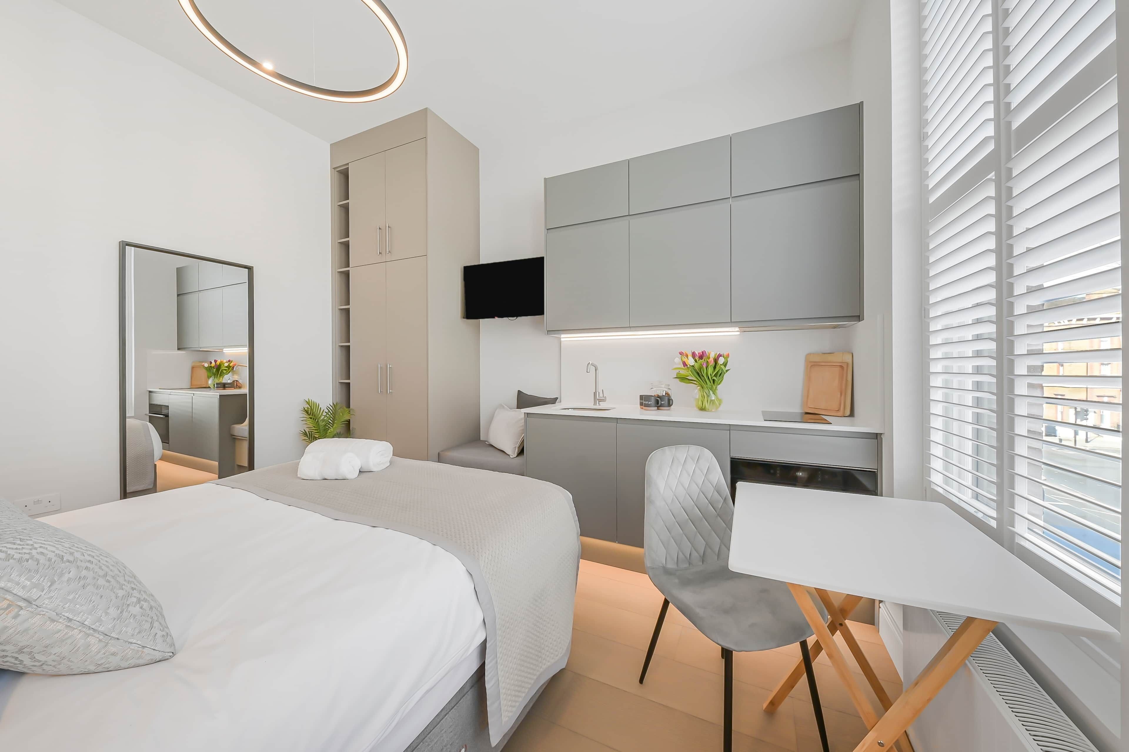 A modern hotel room with a bed, a desk, a TV, and a window with blinds.