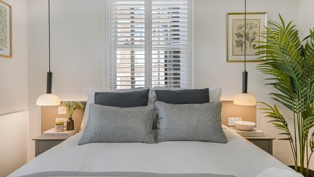 A bedroom with a bed, two bedside tables, a window with blinds, a hanging light fixture, and a potted plant. - Studio 5 @ OneStay Maida Vale (London)