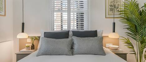 A bedroom with a bed, two bedside tables, a window with blinds, a hanging light fixture, and a potted plant.
