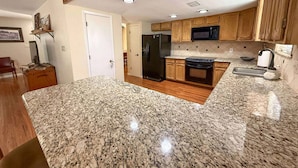 Una cocina con encimeras de granito, armarios de madera, refrigerador, estufa, microondas y pileta.