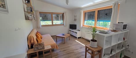 A living room with a sofa, TV, wooden floor, white walls, and a kitchen area with a microwave.