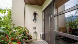 A modern building entrance with a dark door, a potted plant with red flowers, a wall-mounted light fixture, and a small trash can. - Chalet Birkmayer by we rent (Zell am See)