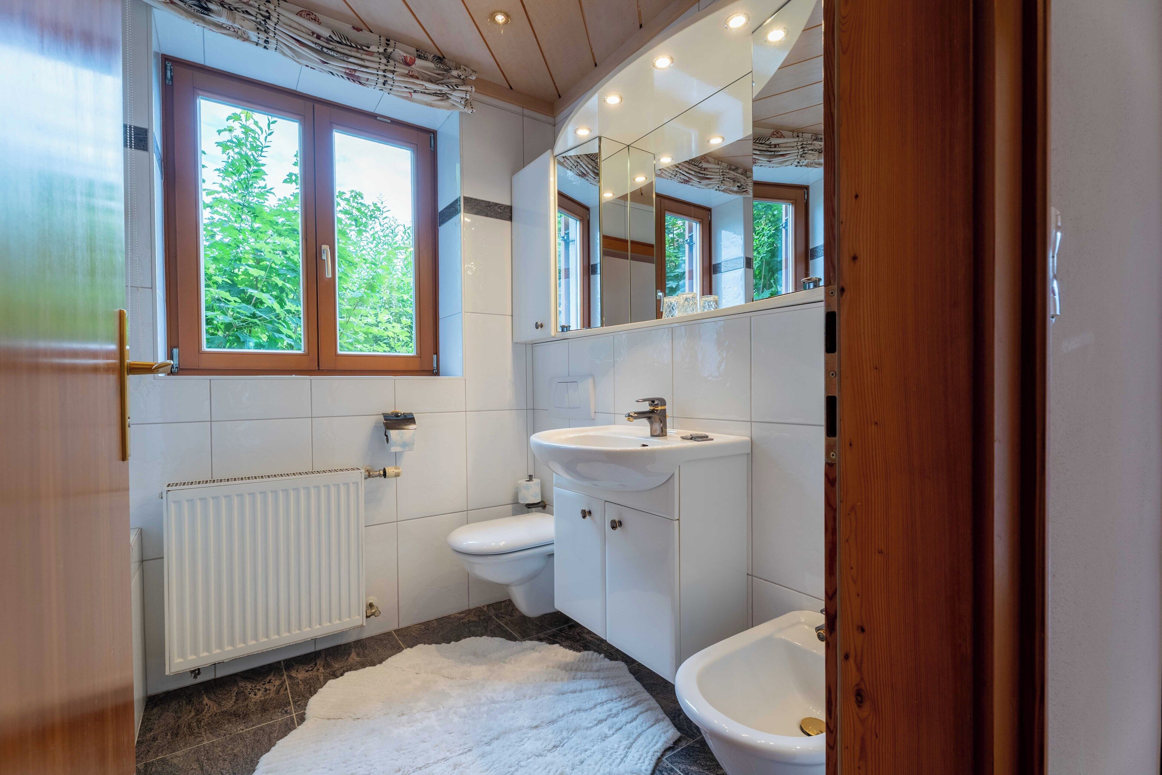 Un baño con lavamanos, inodoro y bidé blancos, un radiador blanco y una ventana con vistas a la vegetación.