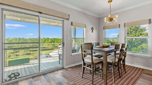A dining area with a table set for six, large windows with blinds, and a view of a wooded area.