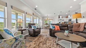 A spacious living room with a large window, leather sofas, a dining area, and a kitchen in the background.