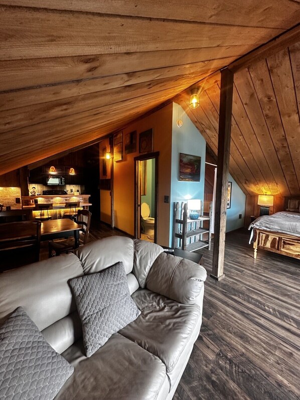 A cozy loft bedroom with a sloped ceiling, a large window, a wooden bed, and a seating area with a sofa and chairs.