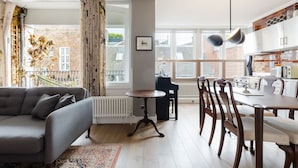 A modern living room with a sofa, a dining table with chairs, a radiator, and a view of a brick building through large windows. - GuestReady - Charming Covent Garden Retreat (London)