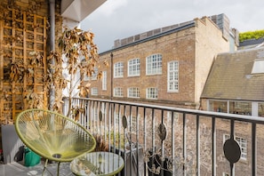 A balcony with a green chair and a glass table, overlooking a brick building with multiple windows.