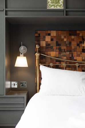 A bed with white bedding, a wall-mounted lamp, a wooden headboard, and a nightstand with a power outlet.