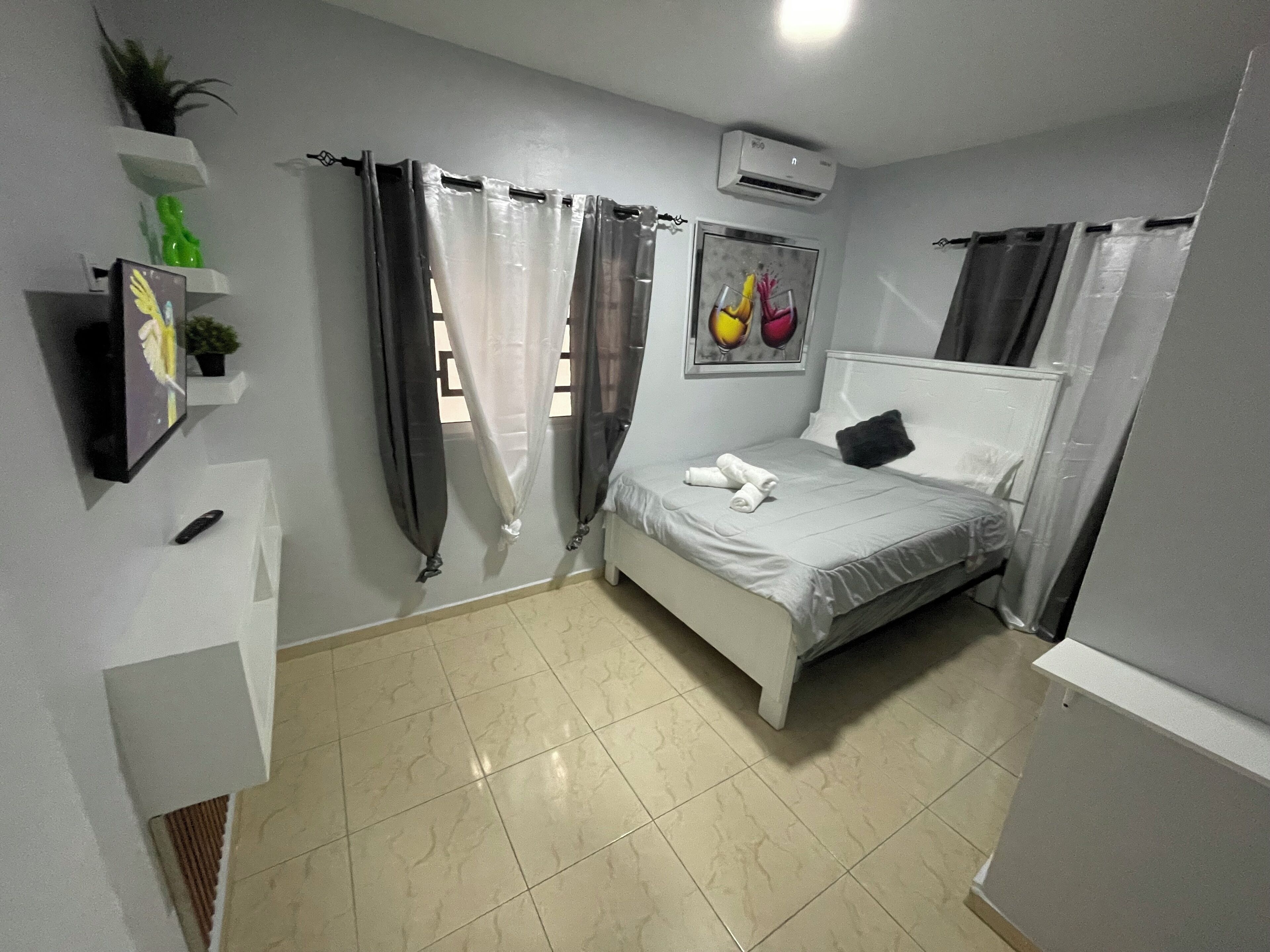 A bedroom with a bed, a white bedside table, a wall-mounted air conditioner, a framed picture, and a potted plant.