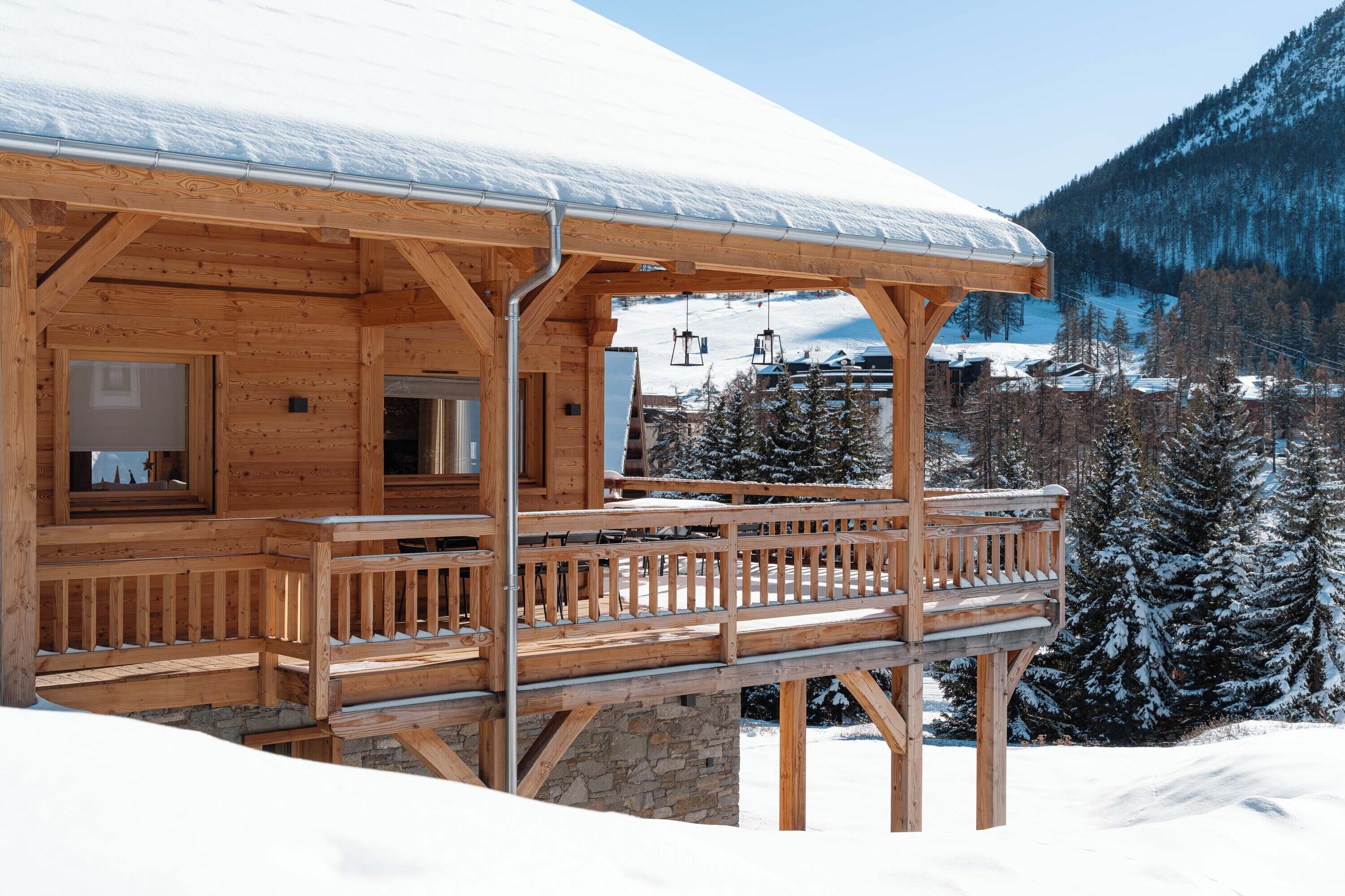 Una cabaña de madera con balcón, rodeada de árboles nevados y montañas.