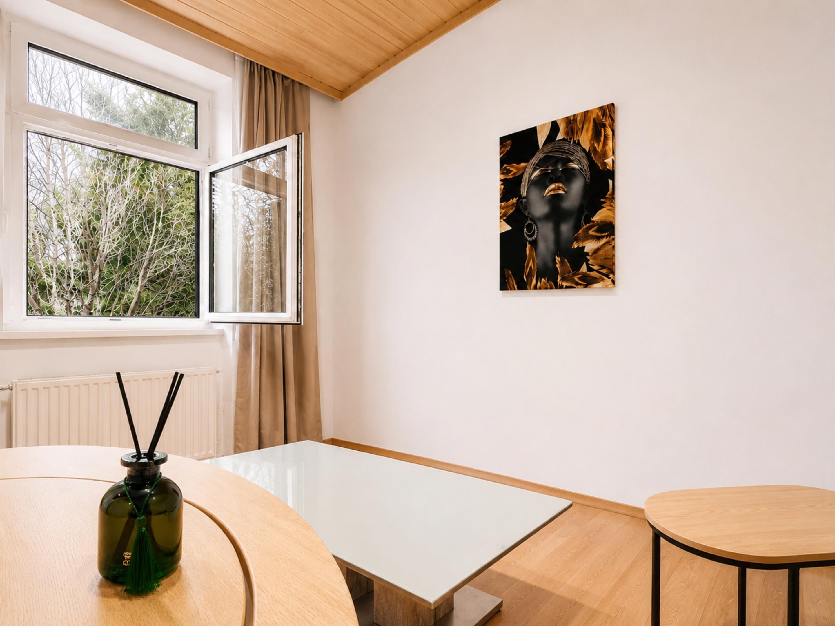 A room with a table, a chair, a window with curtains, and a wall-mounted artwork.