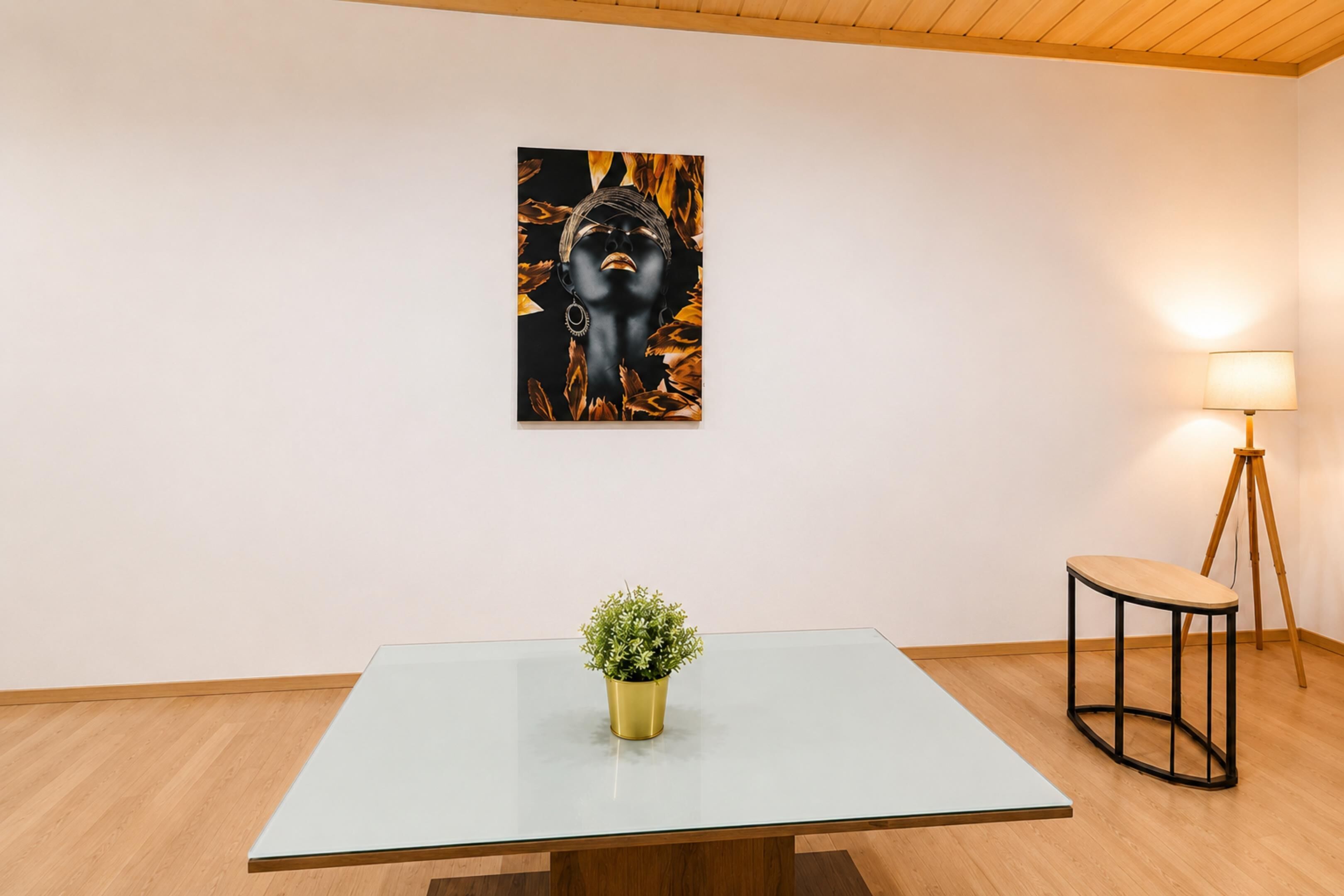 A minimalist room with a glass table, a potted plant, a floor lamp, and a framed artwork on the wall.