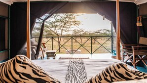 Ein Zelt mit einem Bett, ausgestattet mit Kissen in ZEBRA-Muster, Blick auf die Savanne mit Bäumen und ein Tisch mit Stühlen davor.