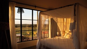 Ein Schlafzimmer mit Himmelbett, großem Fenster mit Blick ins Freie und kleinem Tisch und Stuhl am Fenster.