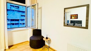 Une pièce avec une grande fenêtre, un pouf noir rond, une petite table ronde avec une bougie et un miroir au mur.