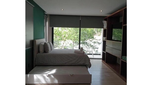 A modern bedroom with a large window, a bed, a built-in bench, and a TV on a wooden shelf. - Practical and Comfortable Loft (Tulum)