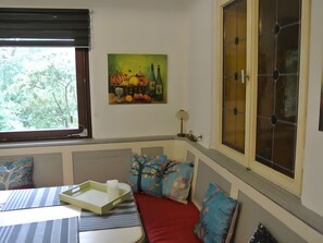 A room with a dining table, a bench, a painting of fruits and bottles, a window with blinds, and a large cabinet with glass doors.