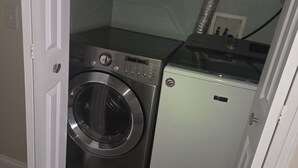 Une petite buanderie équipée d’un lave-linge et d’un sèche-linge à chargement frontal, d’un petit meuble de rangement et d’un conduit d’aération.