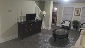 Un salon avec une commode en bois foncé, une table basse ronde en verre et un téléviseur posé sur le dessus. On y trouve un escalier menant à une autre pièce, une chaise blanche et une plante en pot.